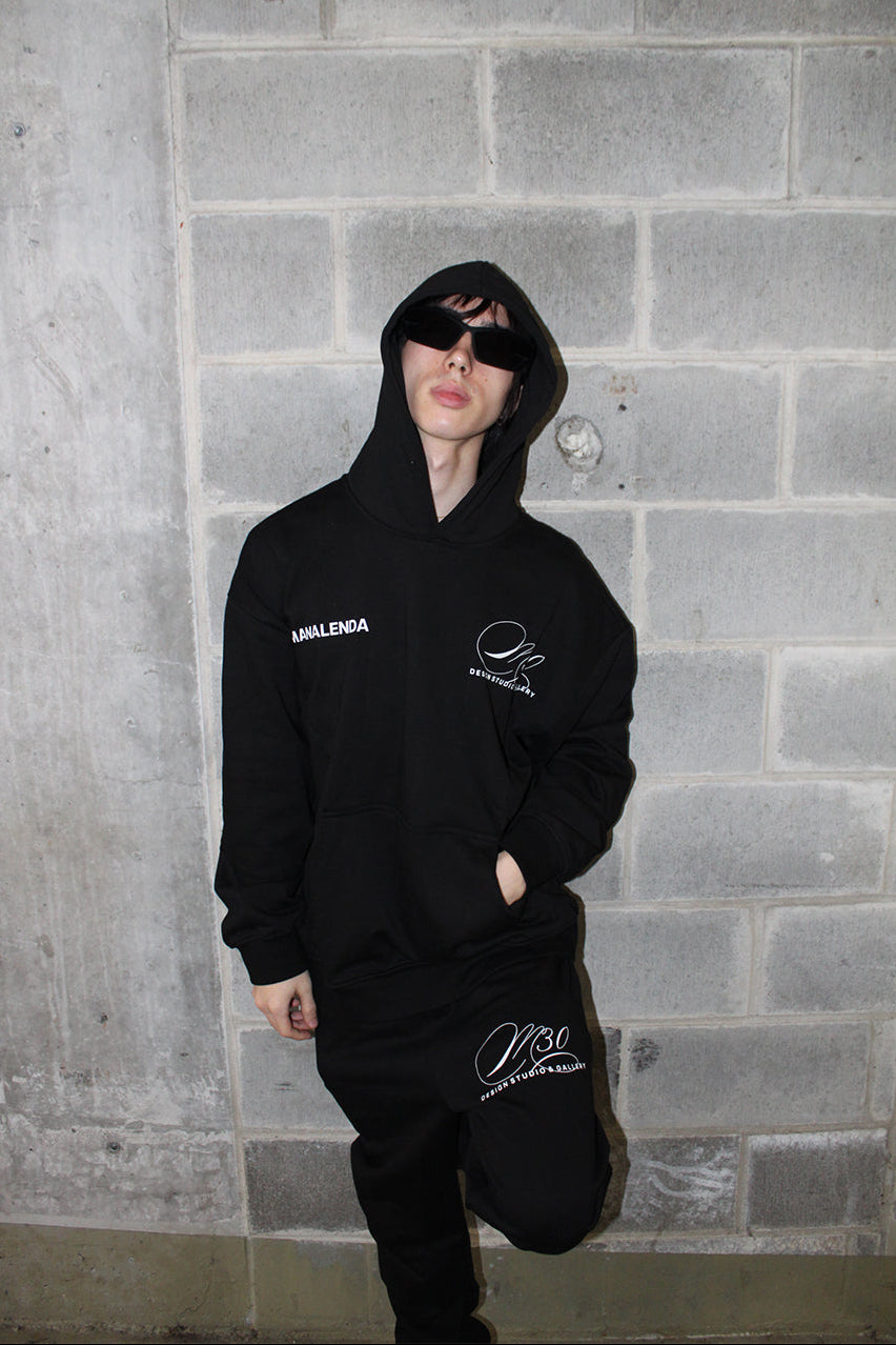 Person wearing a black hoodie with branding against a gray brick wall.